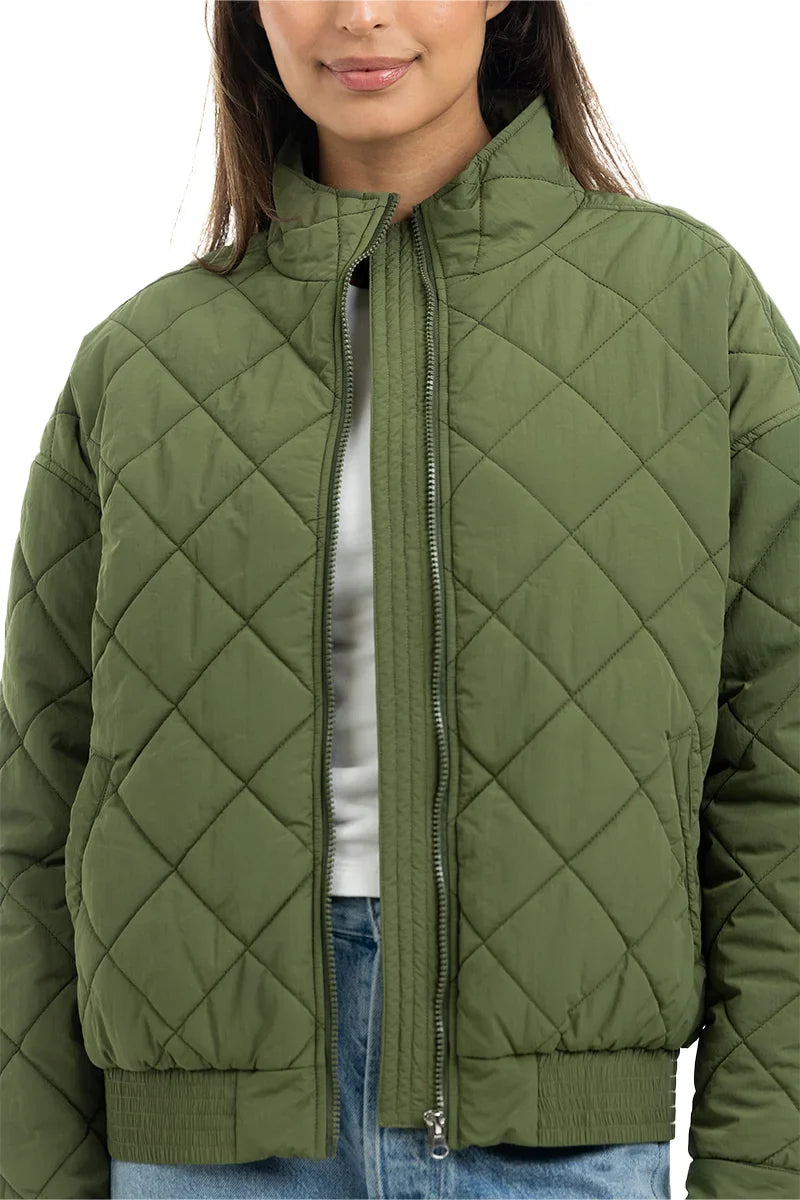 Wearing the Katydid Olive Sporty Diamond Quilted Jacket unzipped over a white top and blue jeans, only the lower half of their face is visible.