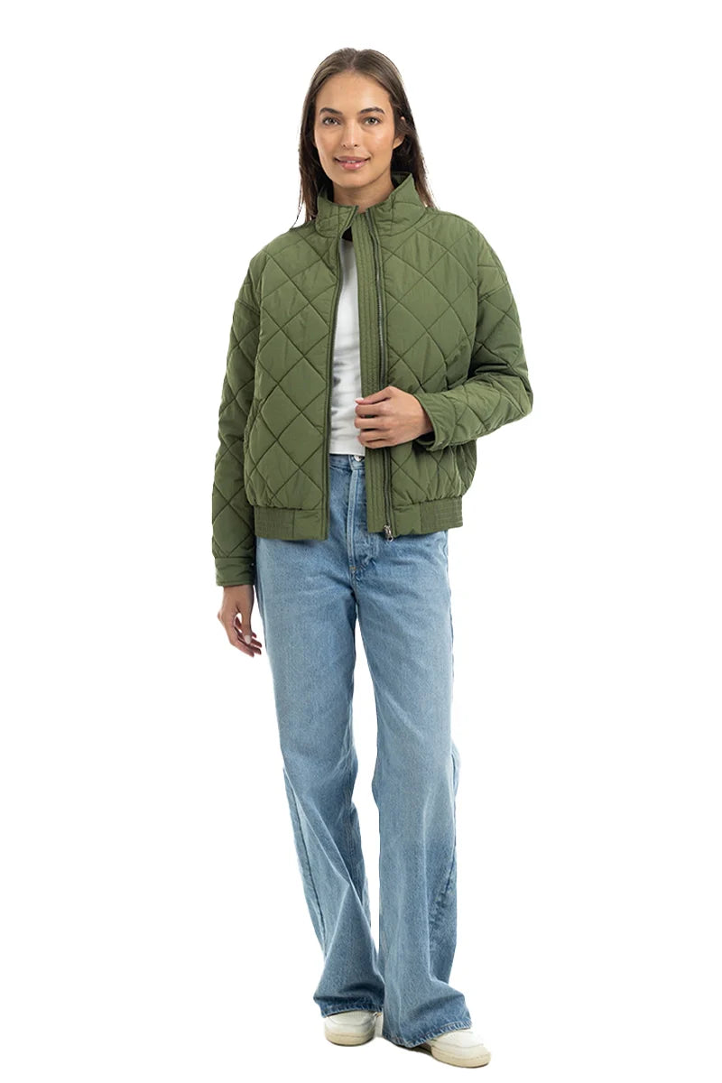 A person stands facing forward, wearing the Katydid Olive Sporty Diamond Quilted Jacket, light blue jeans, and white sneakers, against a plain white background.