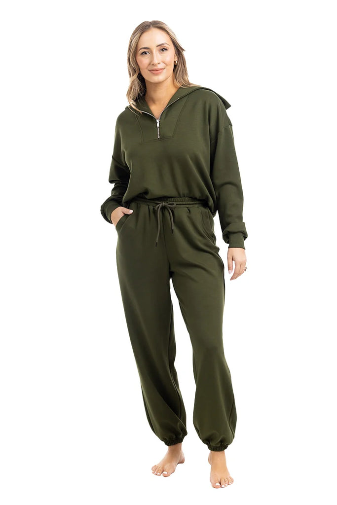 A woman stands barefoot wearing the Katydid Olive Pullover Two-Piece Set, featuring an olive green long-sleeve zip-up hoodie and matching jogger sweatpants. She has her hands in her pockets and smiles at the camera.