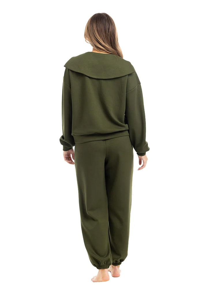 A woman with long hair stands barefoot, facing away, wearing the Katydid Olive Pullover Two-Piece Set—featuring a loose-fitting long-sleeve top and jogger pants—against a plain white background.