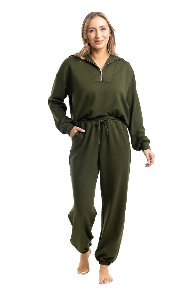 A woman smiles barefoot against a white background, wearing the Katydid Olive Pullover Two-Piece Set—dark green long-sleeve zip-up sweatshirt and matching drawstring jogger pants, with her left hand in her pocket. The set looks both comfortable and casual.