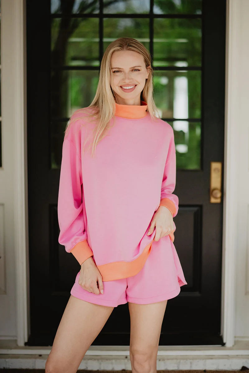 A woman with long blonde hair smiles in front of a black door, wearing the Katydid Pink  Orange Duo Mockneck Sweatset featuring contrast trim at the collar and sleeves, with her hand on her hip.