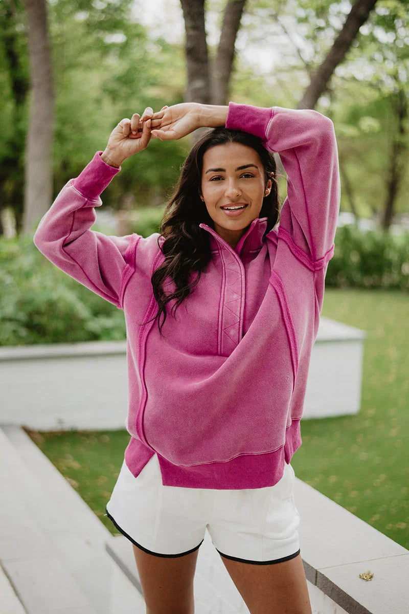 A woman outdoors smiles while stretching her arms overhead, wearing the Katydid Hot Pink Vintage Wash Pullover Womens and white shorts, standing on a path with greenery and trees behind her.