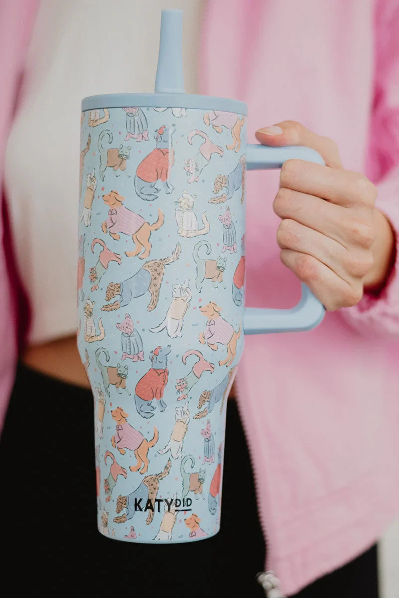 A person in a pink jacket holds the Posh Pets Party Easy Carry Flip Top Tumbler by Katydid, a large light blue reusable cup adorned with colorful cartoon cats and the Katydid logo near the bottom.