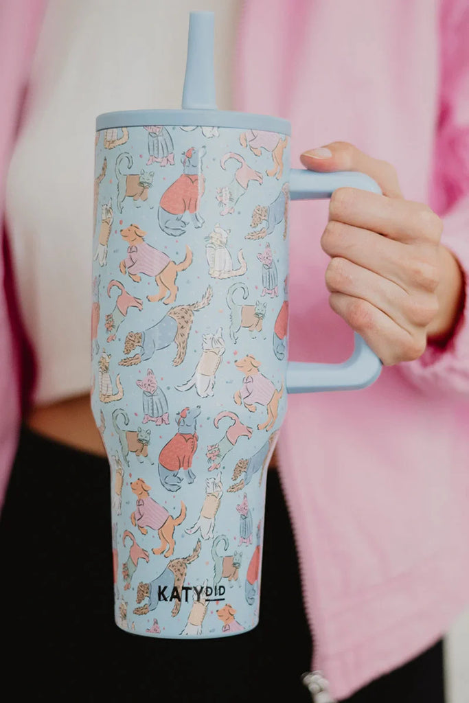 A person in a pink jacket holds the Posh Pets Party Easy Carry Flip Top Tumbler by Katydid, a large light blue reusable cup adorned with colorful cartoon cats and the Katydid logo near the bottom.