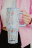 A person in a pink jacket holds the Posh Pets Party Easy Carry Flip Top Tumbler by Katydid, a large light blue reusable cup adorned with colorful cartoon cats and the Katydid logo near the bottom.