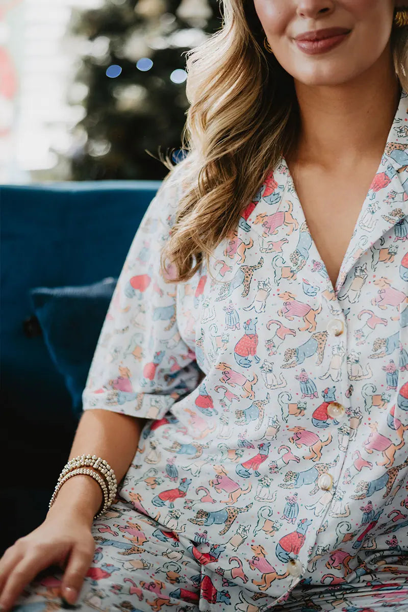 A woman in Katydid’s Posh Pets Preppy Pajama Pants sits on a blue couch, smiling gently. Her wavy blonde hair and gold bracelet complement the pet-themed pants. A blurred Christmas tree glows in the background—ideal for animal lovers.
