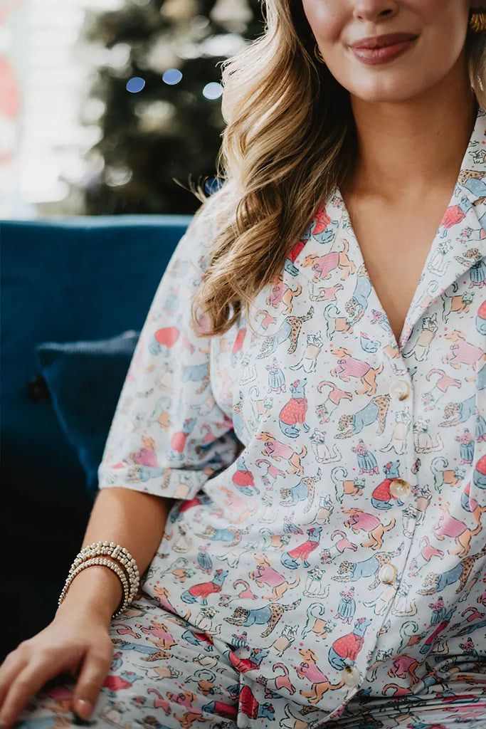 A woman in Katydid’s Posh Pets Preppy Pajama Pants sits on a blue couch, smiling gently. Her wavy blonde hair and gold bracelet complement the pet-themed pants. A blurred Christmas tree glows in the background—ideal for animal lovers.