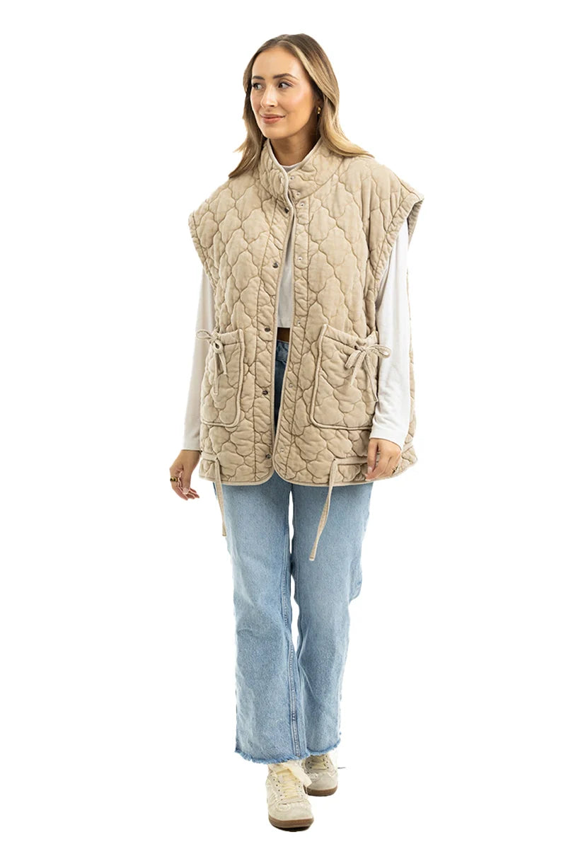 A woman wears the Katydid Beige Oversized Garment Washed Vest over a white long-sleeve shirt with light blue jeans and white sneakers, standing against a crisp white background and smiling slightly.