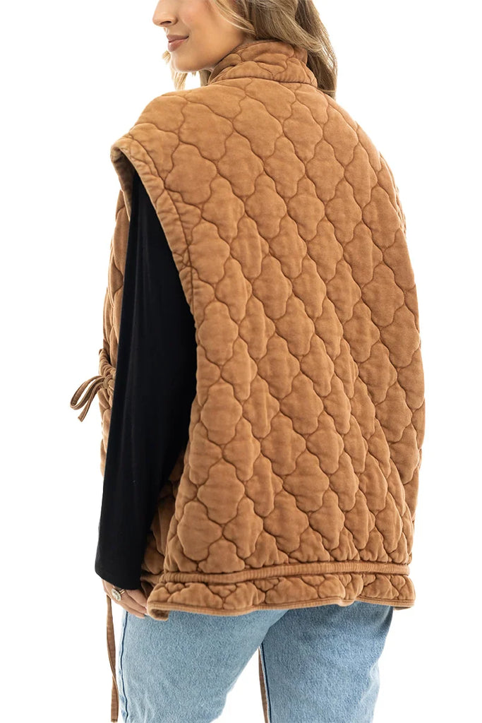 A woman with long hair is seen from the back wearing the Katydid Brown Oversized Womens Quilted Vest over a black top and light blue jeans, with one hand in her pocket.