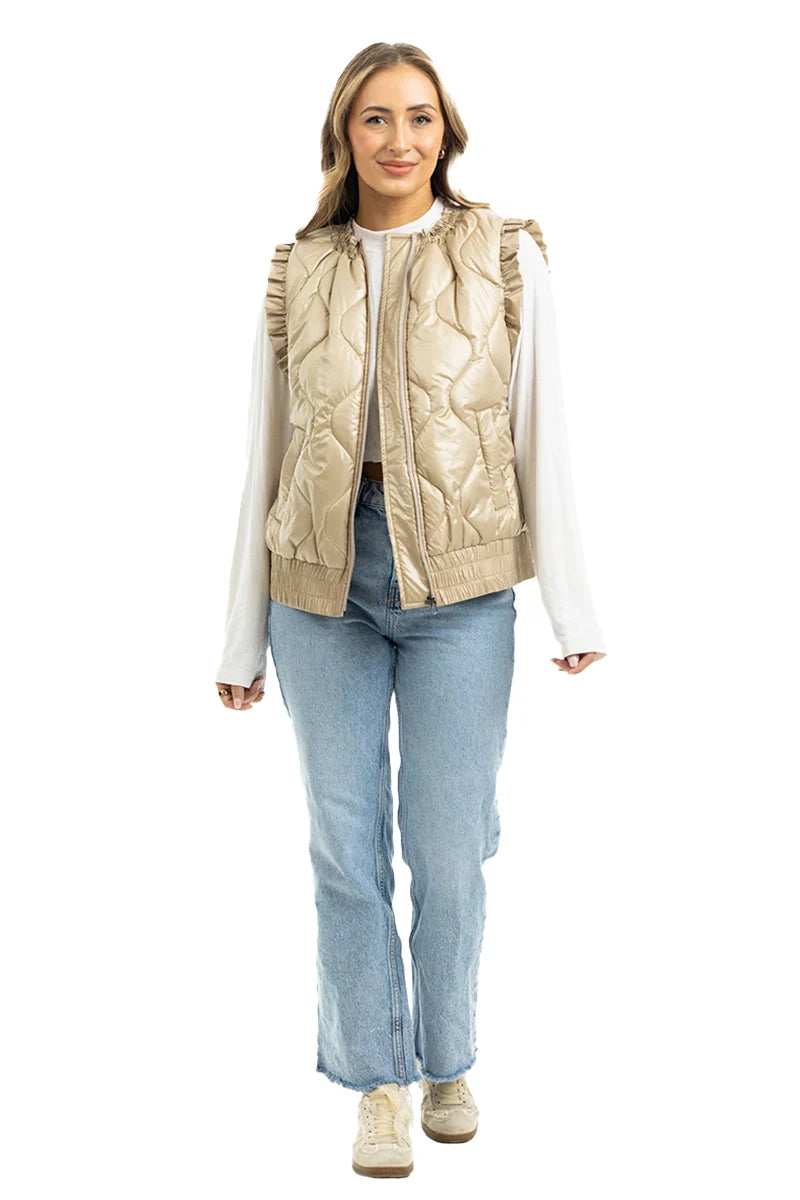 A woman wears the Katydid Soft Tan Metallic Quilted Vest over a white long-sleeve top, with light blue jeans and beige sneakers. She stands facing forward, smiling.