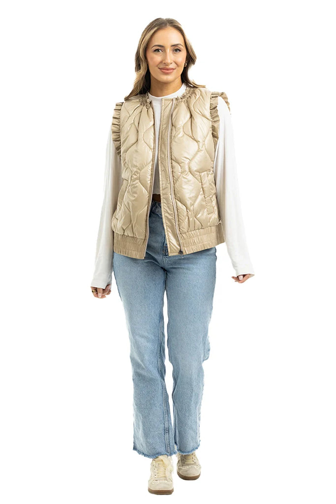A woman wears the Katydid Soft Tan Metallic Quilted Vest over a white long-sleeve top, with light blue jeans and beige sneakers. She stands facing forward, smiling.