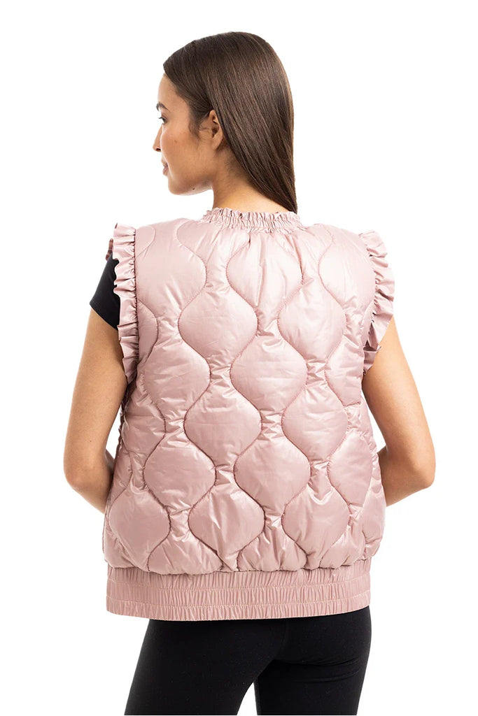 A woman with long brown hair is seen from the back wearing Katydids Blush Metallic Puffy Vest, featuring feminine ruffled shoulders, over a black short-sleeve top and paired with black pants.