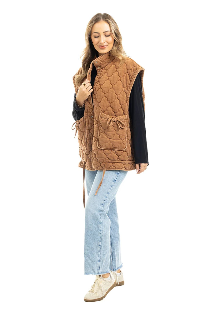 A woman stands smiling against a white background wearing the Katydid Brown Oversized Womens Quilted Vest with large front pockets, paired with a black long-sleeve top, light blue jeans, and white sneakers.