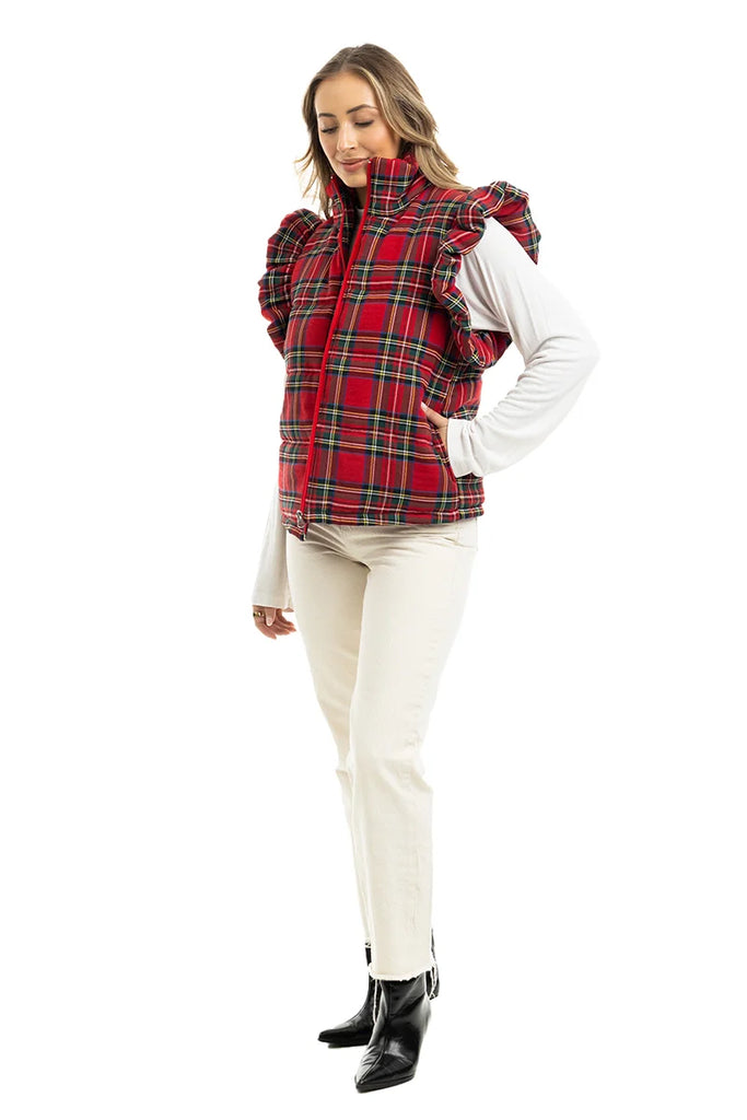 A woman models the Katydid Royal Stewart Plaid Ruffle Puffer Vest, featuring ruffled shoulders, over a white long-sleeve top with white pants and black ankle boots—a festive look against a white background.