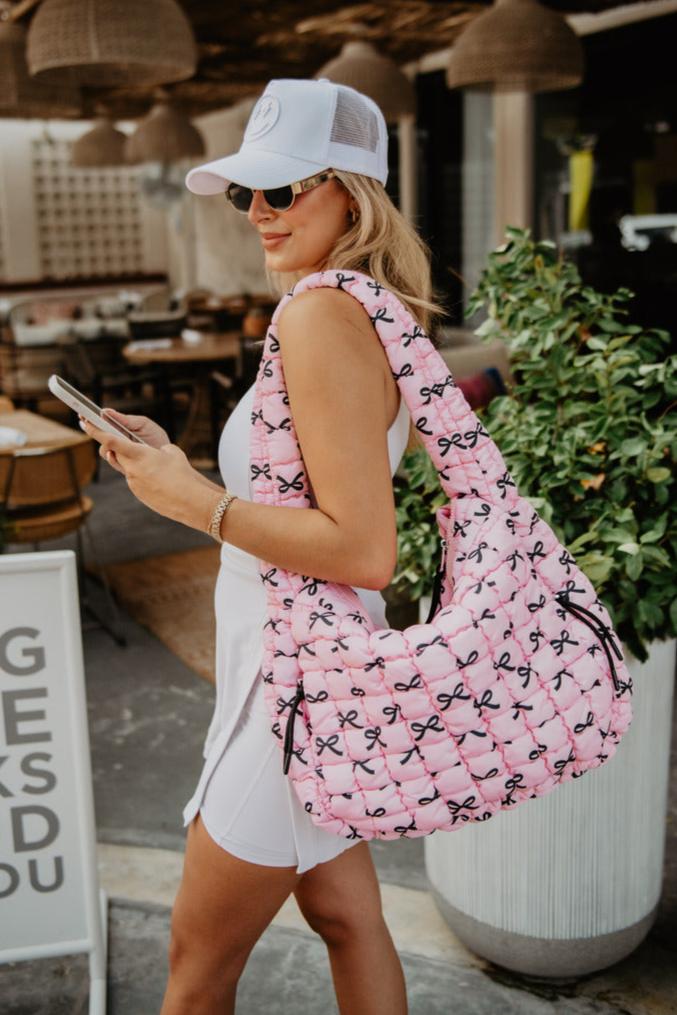 A woman in a white dress and cap holds a phone, wearing sunglasses and a large pink quilted bag with black bows. The scene is casual and summery.