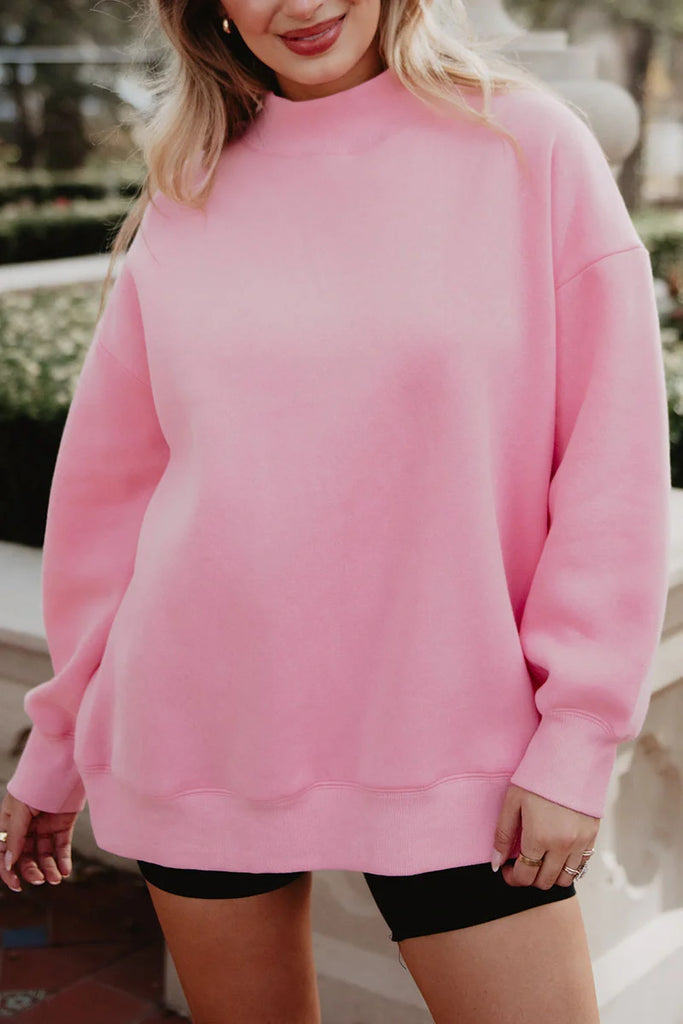 A woman smiles outdoors, wearing the Katydid Pink Essential Mockneck Crew and black shorts, with her hands relaxed by her sides. Only her upper body and part of her legs are visible.