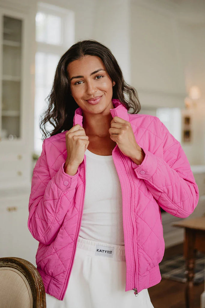 A woman with long dark hair smiles indoors, casually holding the collar of a Katydid Hot Pink Sporty Diamond Quilted Jacket, which she wears over a white top and skirt.