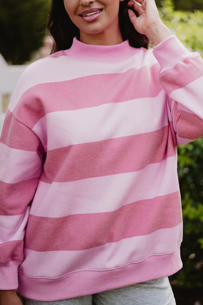 A person in a Katydid Pink and Light Pink Striped Mockneck Pullover stands outdoors, smiling and touching their hair against a blurred green background.