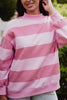 A person in a Katydid Pink and Light Pink Striped Mockneck Pullover stands outdoors, smiling and touching their hair against a blurred green background.