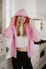 A cheerful woman stands in a bright kitchen, wearing the Katydid Pink Lightweight Casual Hooded Jacket over a white crop top and black pants, holding the hood with both hands.