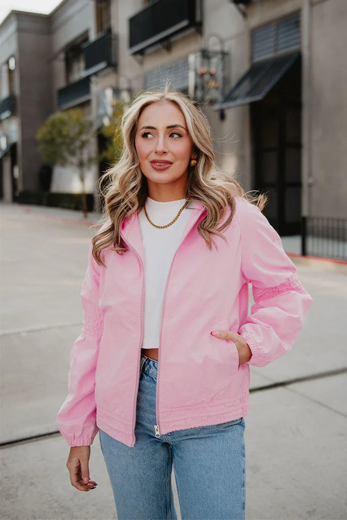 A woman with wavy blonde hair wears the Katydid Pink Lightweight Casual Hooded Jacket over a white crop top and blue jeans, standing outdoors on a city street with buildings in the background.