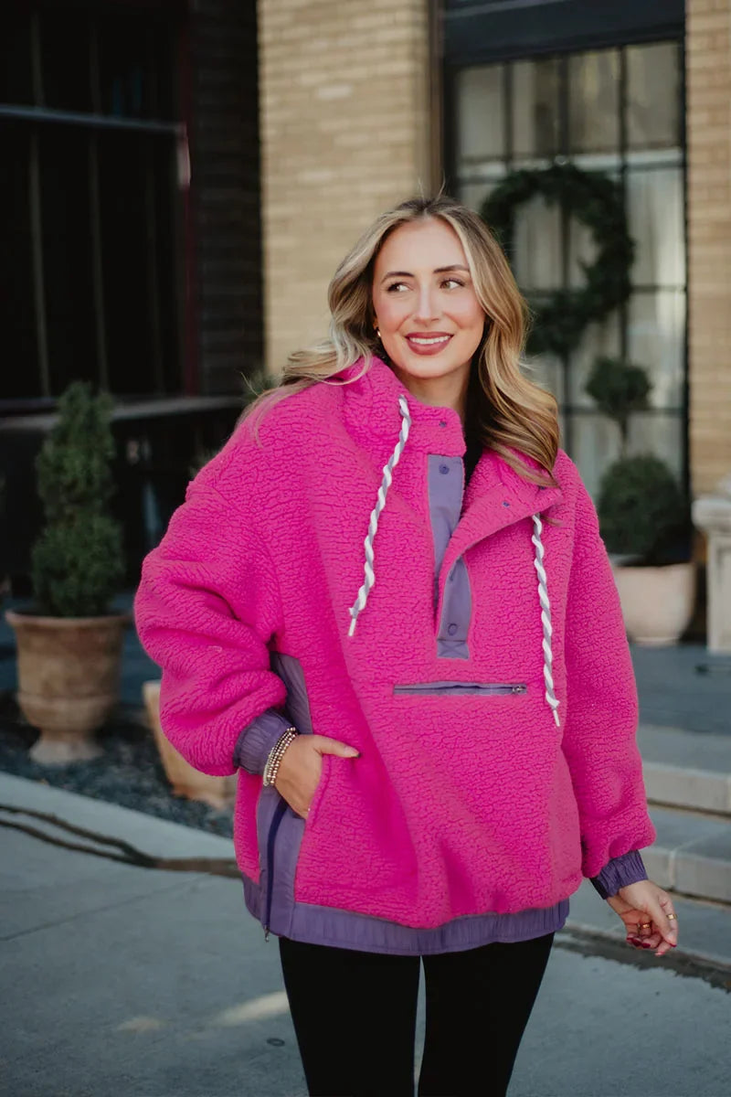 A woman with long, wavy blonde hair stands outdoors by a brick building in potted plants, wearing the Katydid Hot Pink Two-Toned Sherpa Pullover Jacket featuring white drawstrings and a front zipper pocket.