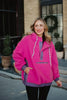 A woman with long, wavy blonde hair stands outdoors by a brick building in potted plants, wearing the Katydid Hot Pink Two-Toned Sherpa Pullover Jacket featuring white drawstrings and a front zipper pocket.