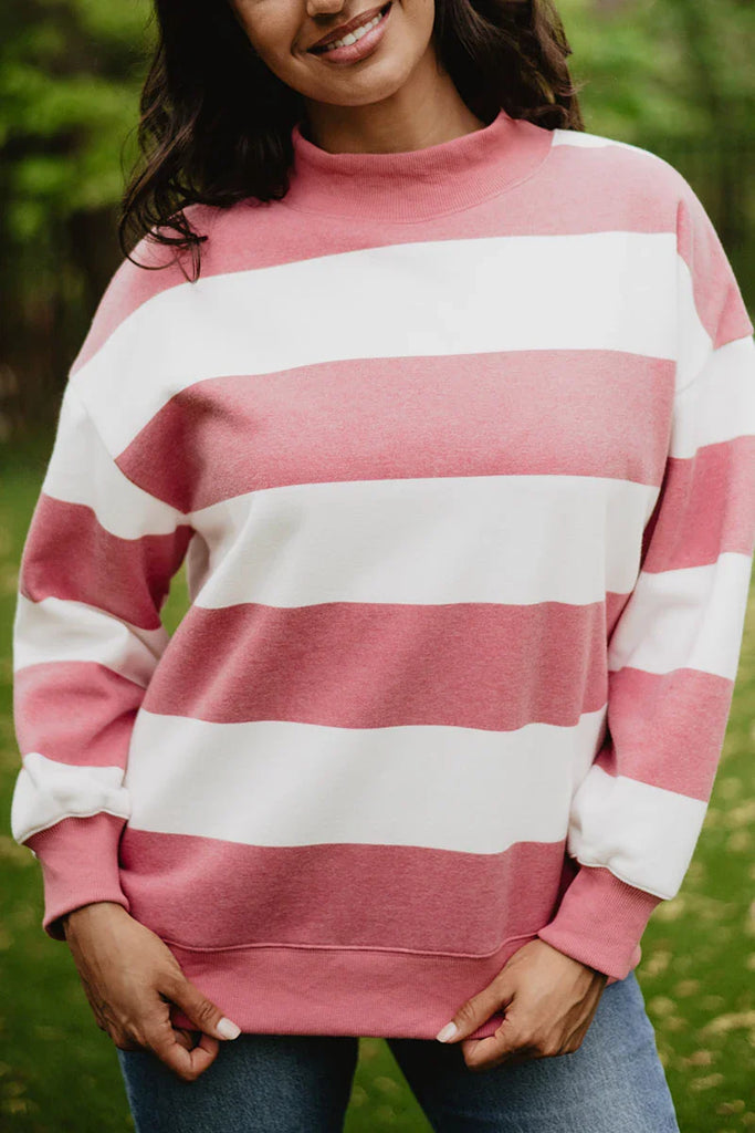 A woman outdoors smiles as she holds the hem of her Katydid Red and White Striped Mockneck Sweatshirt, a cozy long-sleeve top that pairs perfectly with blue jeans. Blurred greenery sets the background for this gameday-ready look.