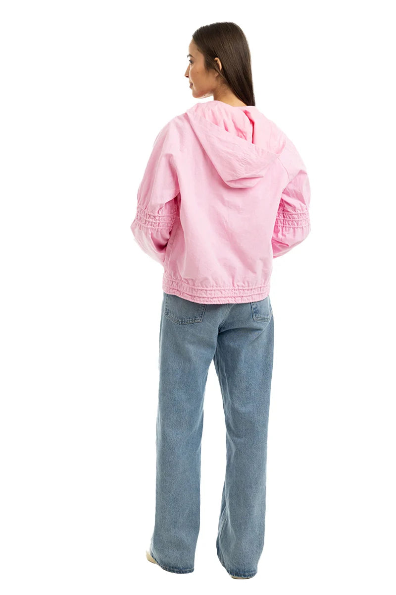 A woman faces away, wearing the Katydid Pink Lightweight Casual Hooded Jacket with gathered sleeves and light blue wide-leg jeans. This women’s jacket is perfect for layering and stands out against a plain white background.