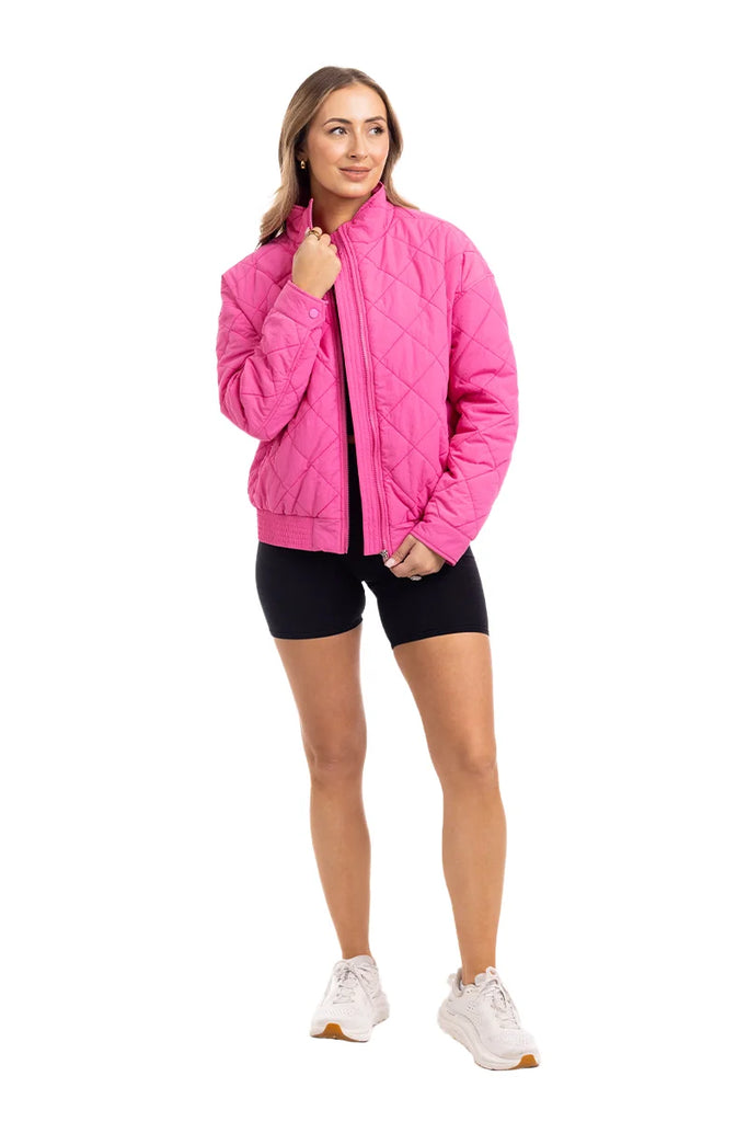 A woman poses against a white background in the Katydid Hot Pink Sporty Diamond Quilted Jacket, paired with black shorts and white sneakers. She holds the jackets collar with one hand and looks to the side, smiling slightly.