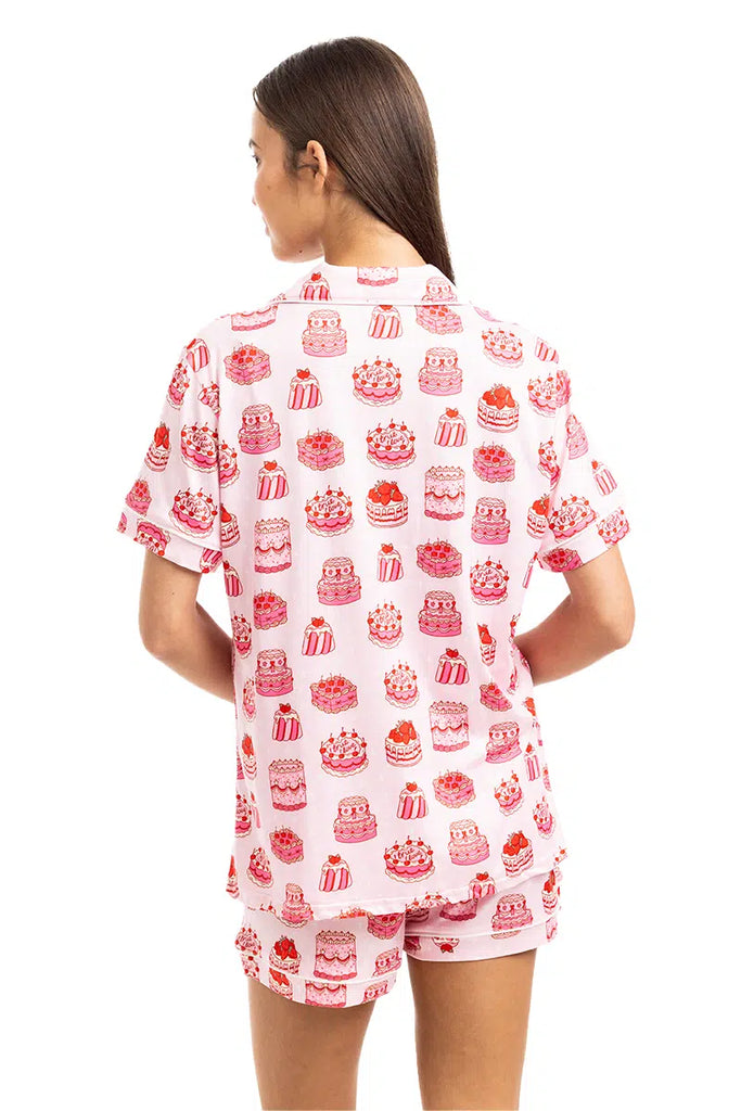 A woman with long brown hair faces away, wearing the Katydid Girly Cakes PJ Shorts Set—a light pink pajama set decorated with playful pink and red cake prints on both the short-sleeve top and matching shorts.