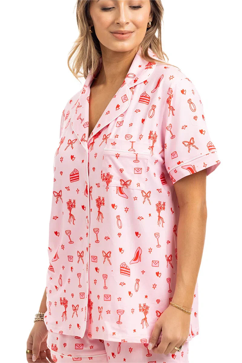 A woman wears the Katydid Literally Just A Girl Icons Short Pajama Set—light pink, short-sleeve button-up with red playful prints of flowers, bows, hearts, heels, and envelopes—smiling slightly for a cute girly pajamas look.