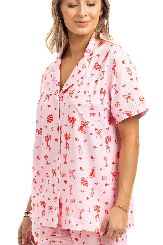 A woman wears the Katydid Literally Just A Girl Icons Short Pajama Set—light pink, short-sleeve button-up with red playful prints of flowers, bows, hearts, heels, and envelopes—smiling slightly for a cute girly pajamas look.