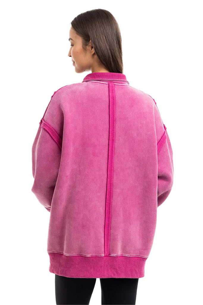 A woman with straight brown hair faces away, modeling the Katydid Hot Pink Vintage Wash Pullover Womens. The oversized pullover features visible seams and ribbed details, set against a plain white background.