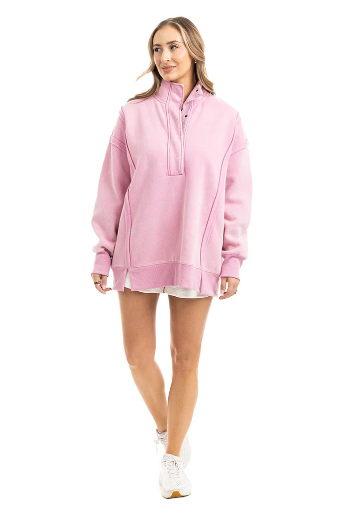 A woman in the Katydid Pink Vintage Wash Pullover for Ladies, styled over a white skirt with white sneakers, stands smiling and looking to the side against a plain white background.