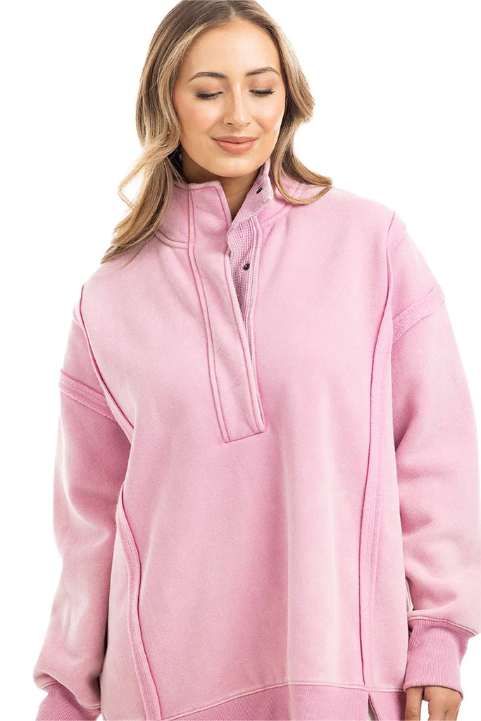 A woman with long, wavy hair wears the Katydid Pink Vintage Wash Pullover for Ladies, featuring an oversized fit and high snap-neck collar. She smiles gently while looking down against a white background.