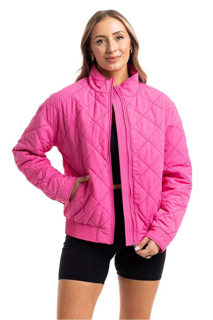 A woman models the Katydid Hot Pink Sporty Diamond Quilted Jacket over a black crop top and shorts, smiling confidently with one hand in her pocket against a white background.