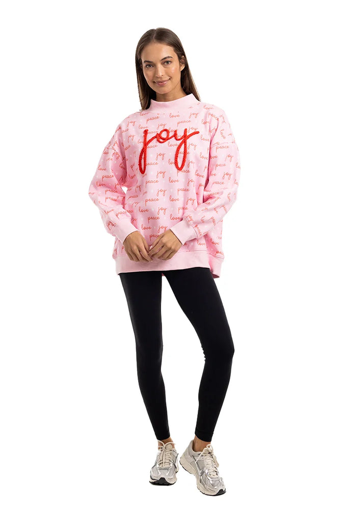 A woman smiles in a Katydid Joy Knit Mockneck Sweatshirt for women, featuring pink fabric with red text and small script details, paired with black leggings and silver athletic shoes against a white background.