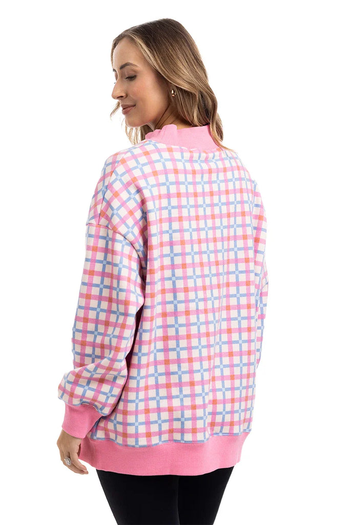 A woman with light brown hair, smiling softly, wears the Katydid Merry  Bright Plaid Mockneck Sweatshirt for women—featuring pink, purple, and white plaid with solid pink trim—paired with black pants.