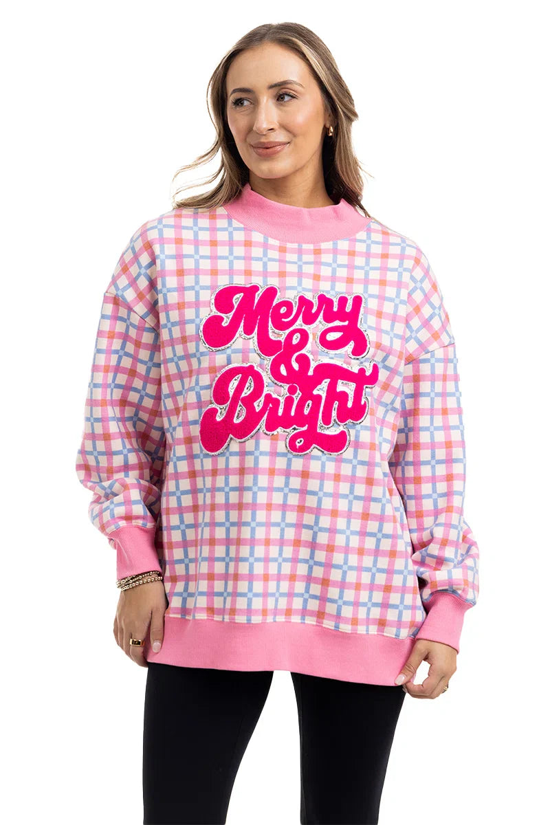 A woman with light brown hair smiles in a Katydid Merry  Bright Plaid Mockneck Sweatshirt for women, featuring pink, blue, and white plaid with bold pink lettering, standing against a white background.