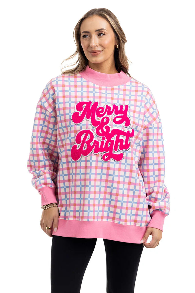 A woman with light brown hair smiles in a Katydid Merry  Bright Plaid Mockneck Sweatshirt for women, featuring pink, blue, and white plaid with bold pink lettering, standing against a white background.