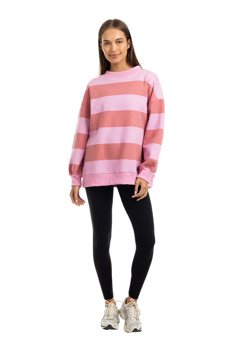 A woman with long brown hair stands facing forward against a white background, wearing the Katydid Pink and Light Pink Striped Mockneck Pullover, black leggings, and white sneakers, smiling gently with her arms relaxed at her sides.