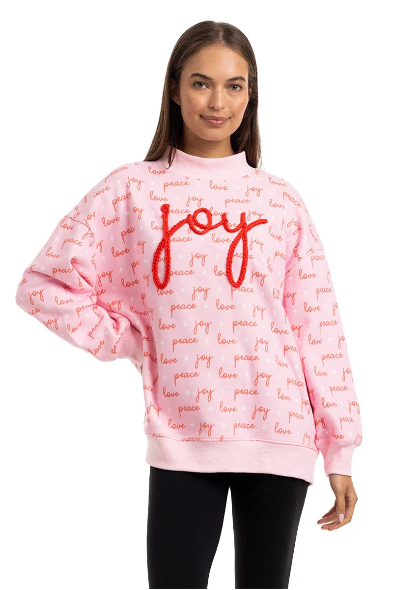 A woman wears the Katydid Joy Knit Mockneck Sweatshirt, a light pink Christmas top with red text that repeats joy, love, and peace and features large, textured red joy lettering on the front.