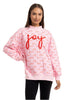 A woman wears the Katydid Joy Knit Mockneck Sweatshirt, a light pink Christmas top with red text that repeats joy, love, and peace and features large, textured red joy lettering on the front.
