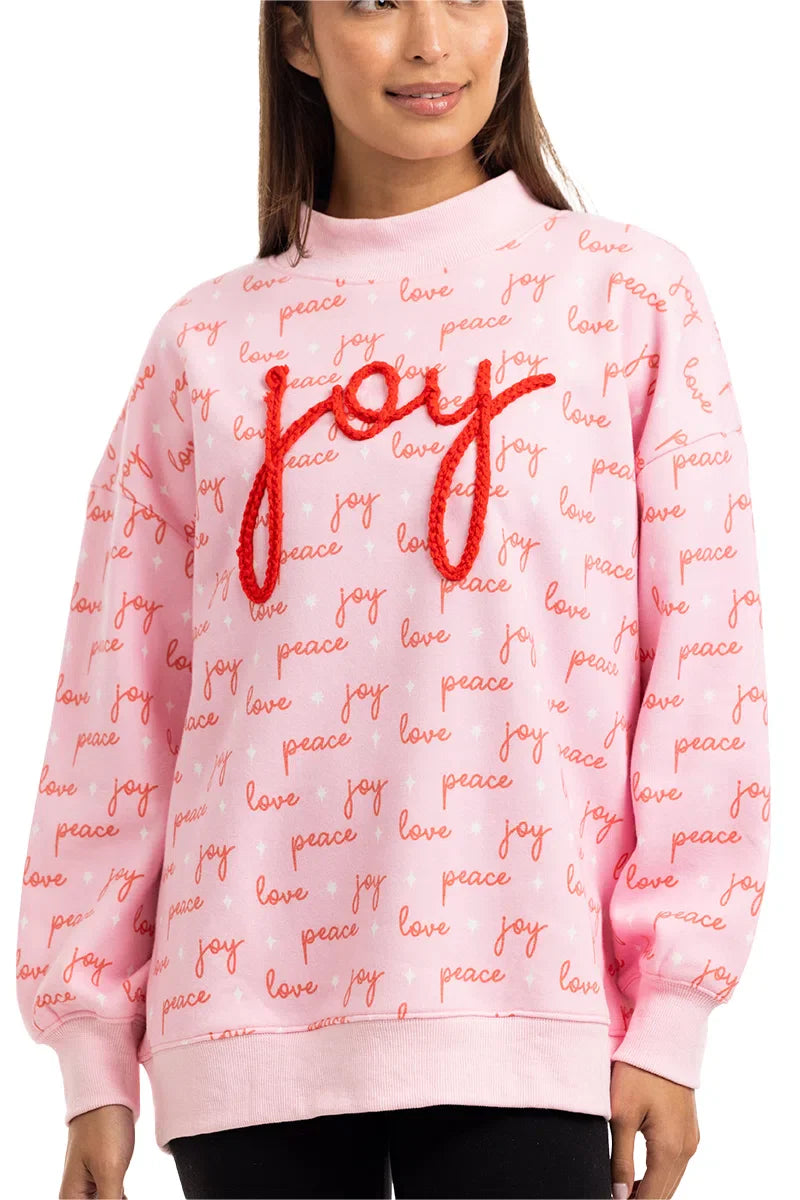 A woman wears the Katydid Joy Knit Mockneck Sweatshirt, a pink pullover for women featuring festive red joy, love, peace patterns and the word joy boldly embroidered in large red letters across the chest.