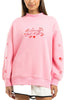 A woman wears the Katydid Literally Just A Girl Mockneck Pullover, a cute light pink sweatshirt with red text and small embroidered flower, heart, and diamond details, paired stylishly with a white skirt.