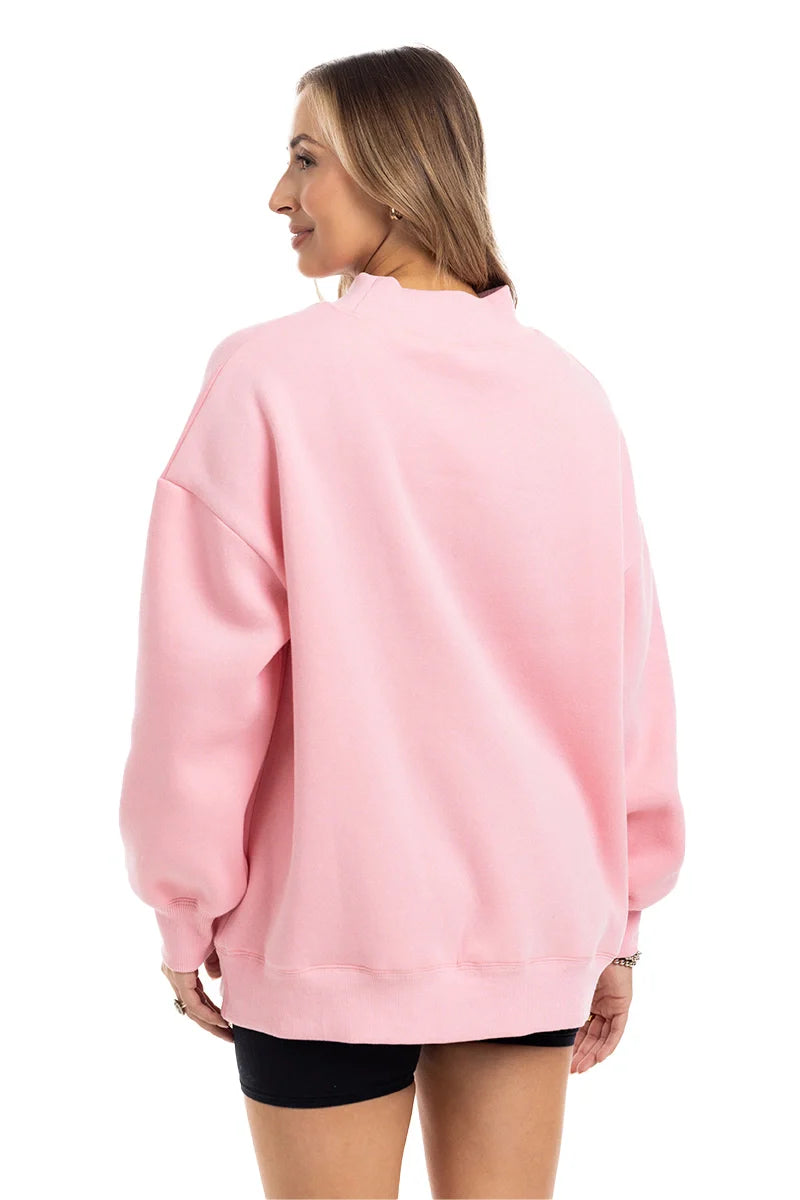 A woman with straight, light brown hair wears the Katydid Pink Essential Mockneck Crew and black shorts, standing against a white background and smiling slightly, with her hands relaxed at her sides.