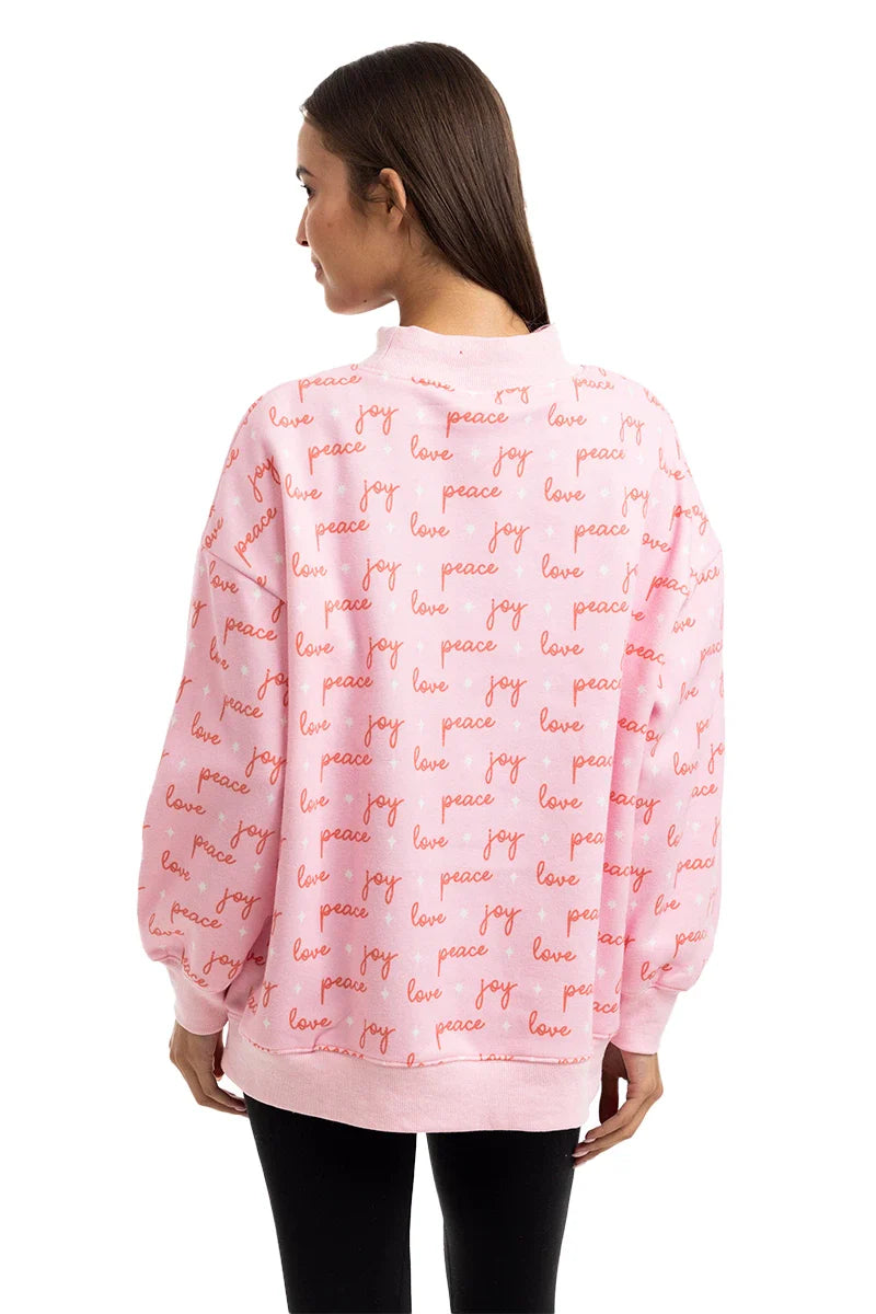 A woman with straight brown hair, seen from behind, wears a loose-fitting Katydid Joy Knit Mockneck Sweatshirt Women in pink with red script reading peace, love, and joy all over. She pairs this festive pullover with black leggings.
