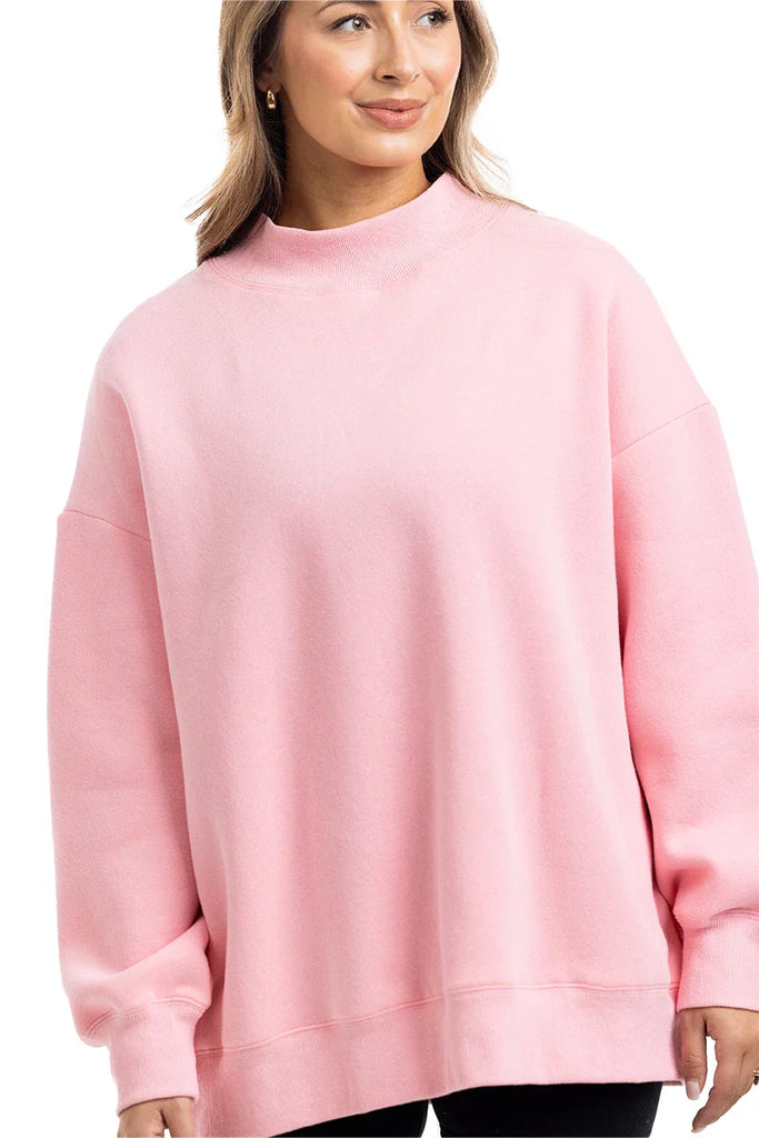A woman models the Katydid Pink Essential Mockneck Crew, a cozy light pink sweatshirt with long sleeves and a high neckline, against a plain white background.